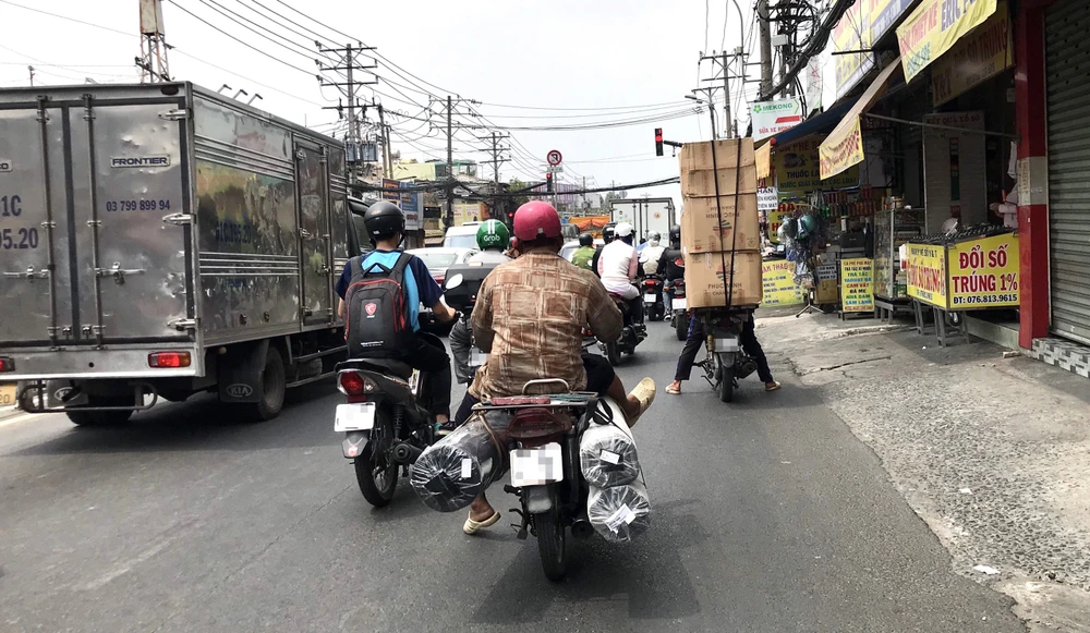 Không có chỗ gác chân, người chở hàng đành gác lên những món hàng mà mình đang chở. Phía trước là một thanh niên khác chở những thùng hàng chất cao. Ảnh: TRẦN MINH