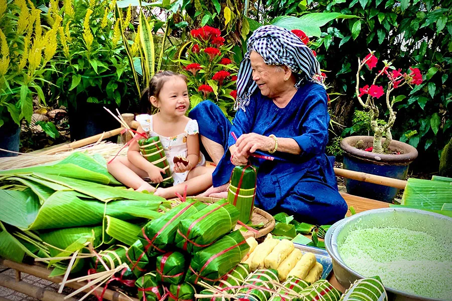 Cụ Nguyễn Thị Ích cẩn thận siết chặt mối dây lạt buộc đòn bánh tét, đứa cháu ngoan ngoãn ngồi cạnh phụ bà lấy dây. Ảnh: TÚ NGÂN