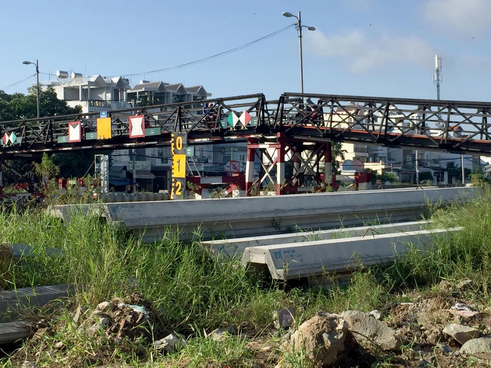 Cây cầu tạm bắc ngang dòng kênh đen đặc rất đông người dân đi lại vào giờ cao điểm, các trụ bê tông của dự án thì nằm ngổn ngang phơi nắng mưa.