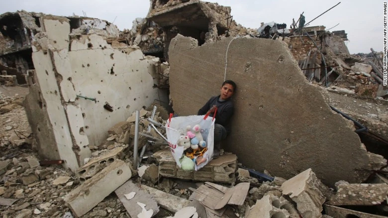 Cậu bé Syria cùng hành lý sơ tán ngồi trên đống đổ nát trước đó là nhà của mình ở quận Al-Arkoub (Aleppo) ngày 17-12. Ảnh: CNN 