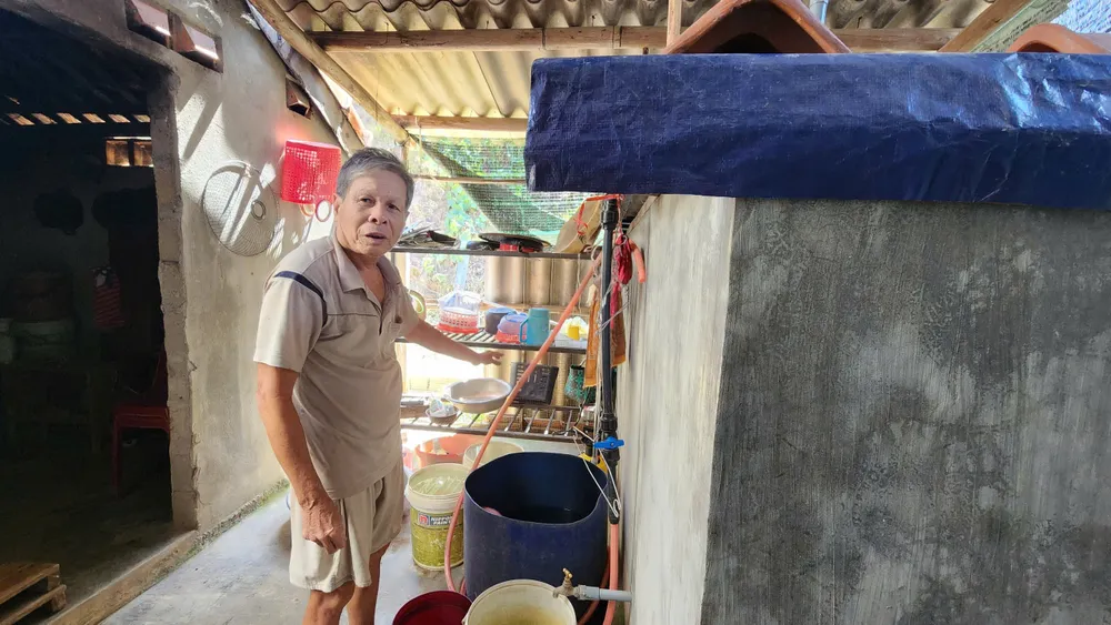 Ông Nguyễn Phơ bên bể chứa nước nhiễm phèn mà gia đình phải sử dụng hàng ngày, nguồn nước này được dẫn từ bể chứa nước ở nghĩa trang. Ảnh: NGUYỄN DO nguoi-dan-hai-hung-vi-gan-10-nam-su-dung-nuoc-o-nghia-trang (4).jpg