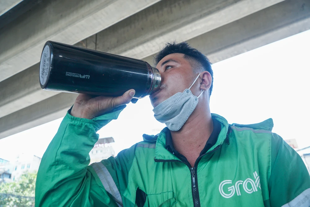 Cạnh đó, một tài xế xe ôm công nghệ vừa hoàn thành việc trả khách, tranh thủ đỗ vào gầm cầu để uống nước lấy lại sức khỏe giữa thời tiết oi bức. Người lao động tự do thường tận dụng gầm cầu, bóng cây là nơi nghỉ ngơi sau những giờ làm việc vất vả.