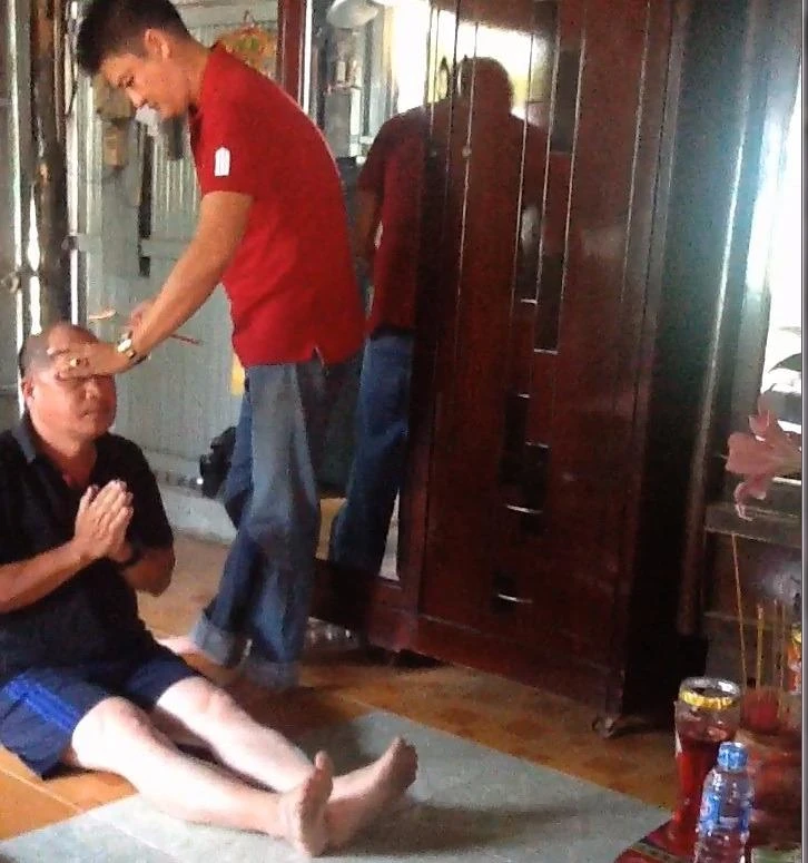 Người đàn ông áo đỏ vừa ấn tay vô trán và thái dương, vừa quơ bó nhang cháy đỏ 