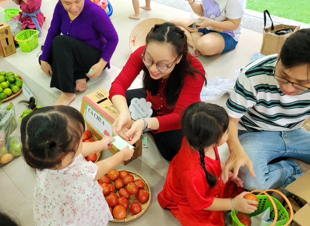 Bé gái lễ phép đưa phiếu bằng hai tay, bạn bên cạnh đang chọn mua cà chua.