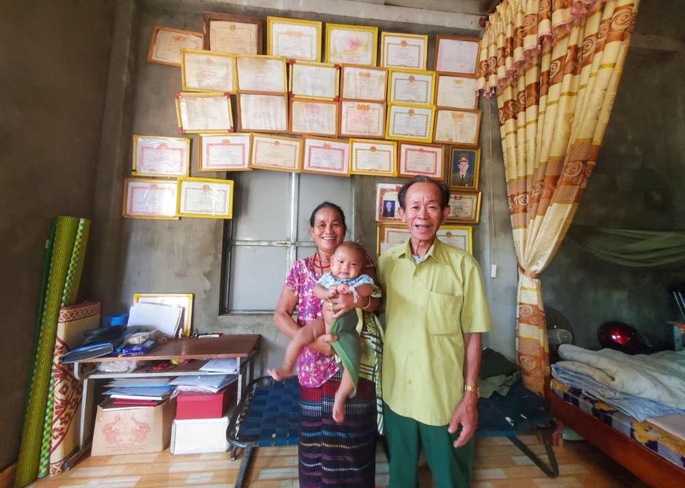 Vợ chồng ông Hồ Hơn cùng đứa chắt đầu trong căn nhà nhỏ cấp 4 và những tấm giấy khen ghi nhận sự đóng góp của ông. Ảnh: BẢO THIÊN