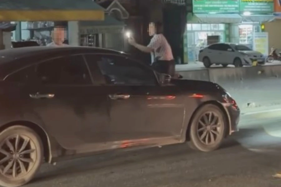 Người phụ nữ ngồi trên nắp capo xe hơi trên quốc lộ 1A và chiếc xe di chuyển.