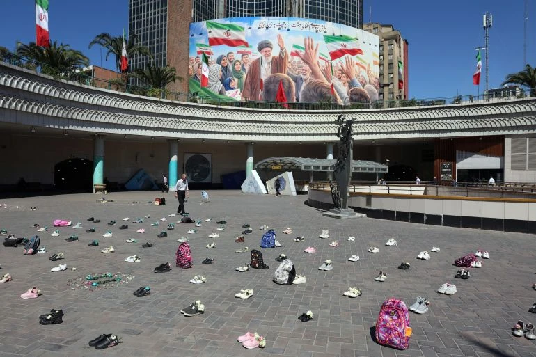 Người dân Iran đi bộ trước một tấm áp phích khổng lồ về cố Lãnh tụ Tối cao Ayatollah Ali Khamenei. Bên cạnh đó là những vật dụng mang tính biểu tượng của các nữ sinh một trường tiểu học ở TP Minab (tỉnh Hormozgan, miền nam Iran) thiệt mạng hồi đầu xung đột, được đặt trên mặt đất tại Quảng trường Valiasr ở Tehran. Ảnh: Abedin Taherkenareh/EPA