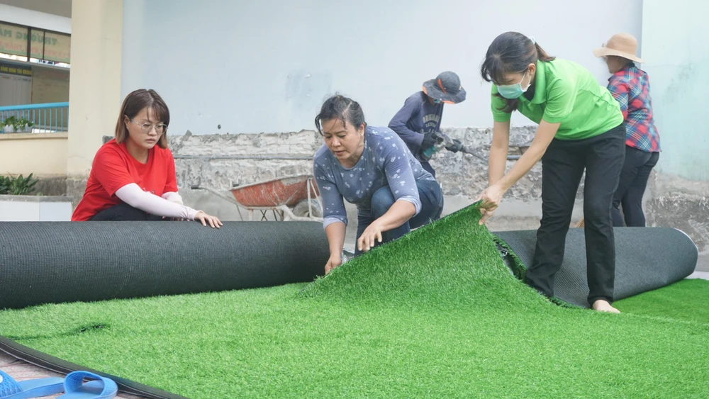 Trước thềm năm học mới, nhà trường đầu tư thêm thảm cỏ để tạo sân bóng nhân tạo cho trẻ vui chơi. Bà Nguyễn Thị Kim Oanh, Hiệu trưởng nhà trường (ở giữa) cùng đội ngũ nhân viên đang cắt ghép, sắp xếp lại khu vực sân cỏ.