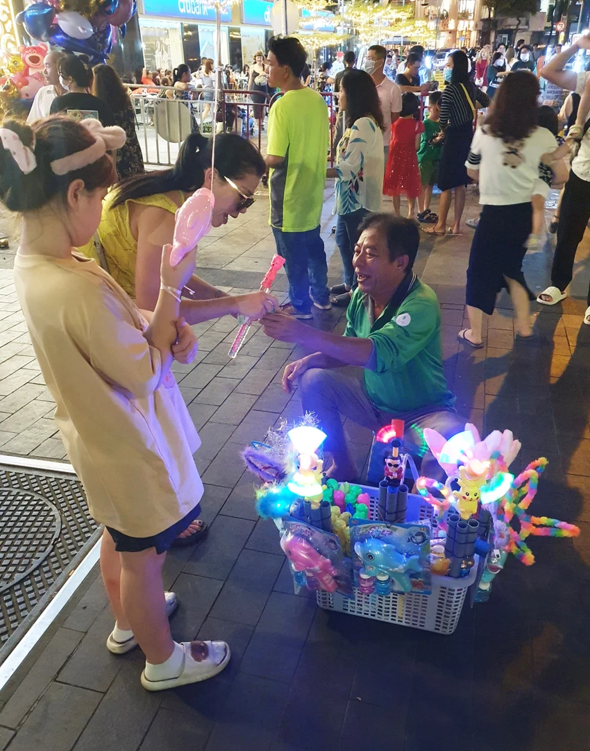 Càng gần đến thời khắc giao thừa, người dân TP.HCM đổ về khu vực trung tâm ngày một đông hơn. Hòa lẫn vào dòng người cũng có những số phận vất vả tranh thủ bán thêm chút ít. Chú Hiền, người đang cười vui bán hàng cho khách trên đường Nguyễn Huệ cho biết nhà ở huyện Nhà Bè, sáng thì chạy grab, tối ra khu vực này để mong bán được nhiều hơn. Càng gần đến thời khắc giao thừa, người dân TP.HCM đổ về khu vực trung tâm ngày một đông hơn. Hòa lẫn vào dòng người cũng có những số phận vất vả tranh thủ bán thêm chút ít. Chú Hiền, người đang cười vui bán hàng cho khách trên đường Nguyễn Huệ cho biết nhà ở huyện Nhà Bè, sáng thì chạy grab, tối ra khu vực này để mong bán được nhiều hơn.